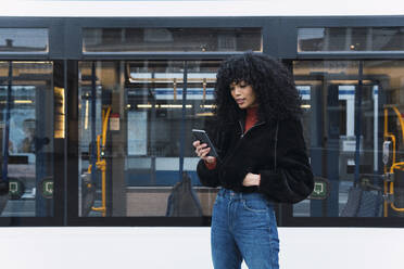 Modische junge Frau mit Smartphone vor einer Straßenbahn - PNAF02957