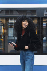 Lächelnde junge Frau mit Smartphone vor einer Straßenbahn - PNAF02958