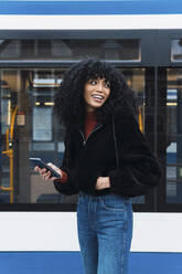 Smiling young woman with smart phone in front of tram - PNAF02958