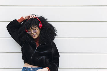 Happy young fashionable woman listening music through headphones in front of white wall - PNAF02962