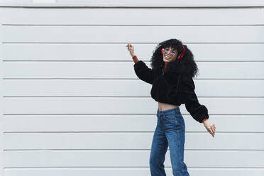 Junge modische Frau hört Musik über Kopfhörer und tanzt vor einer weißen Wand - PNAF02964