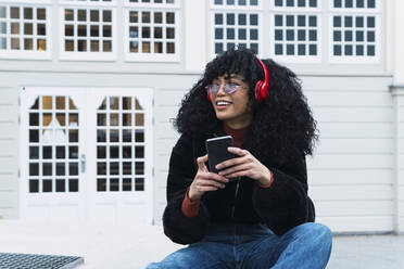 Fashionable young woman sitting with smart phone in front of building - PNAF02965