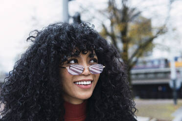 Straßenmarkierung, die sich auf der Sonnenbrille einer lächelnden jungen Frau mit lockigem schwarzem Haar spiegelt - PNAF02970