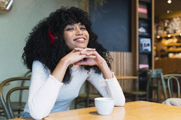 Lächelnde junge Frau mit gefalteten Händen auf dem Kinn hört Musik in einem Cafe - PNAF02980