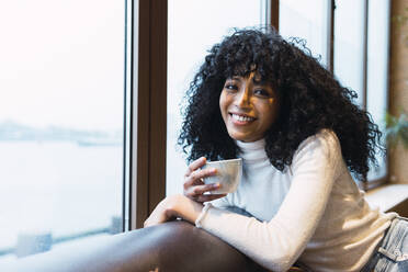 Smiling curly haired woman with coffee cup leaning on sofa at cafe - PNAF02982