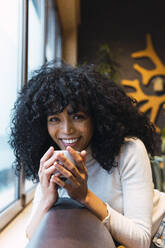 Lächelnde, lockige, schwarzhaarige Frau mit Kaffeetasse, die sich im Café auf das Sofa lehnt - PNAF02983