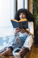 Nachdenkliche junge Frau sitzt mit einem Roman am Fenster eines Cafés - PNAF02989