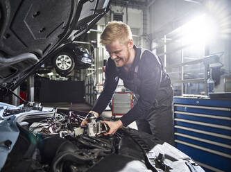 Lächelnder junger blonder Mechaniker, der einen Automotor in einer Autowerkstatt repariert - CVF01880