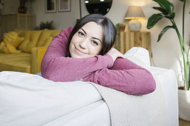 Smiling beautiful woman leaning on sofa at home - XLGF02577