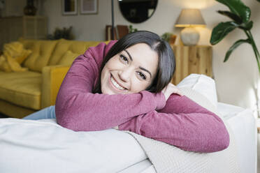 Happy young woman leaning on sofa in living room - XLGF02578