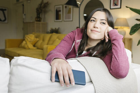 Nachdenkliche Frau mit Smartphone auf dem Sofa im Wohnzimmer sitzend - XLGF02582