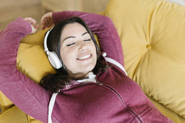 Happy woman with eyes closed listening music through wireless headphones sofa at home - XLGF02587