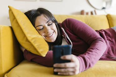 Smiling woman surfing net through smart phone on sofa at home - XLGF02591