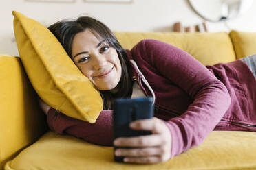 Smiling woman with mobile phone lying on sofa at home - XLGF02592