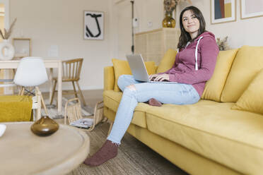 Lächelnde Frau mit Laptop auf dem Sofa im Wohnzimmer sitzend - XLGF02596