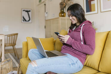 Smiling young woman sitting with laptop text messaging through smart phone on sofa - XLGF02598