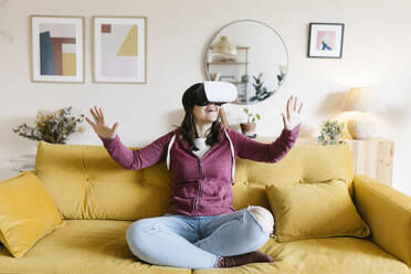 Glückliche Frau mit Virtual-Reality-Headset gestikuliert auf dem Sofa zu Hause - XLGF02615