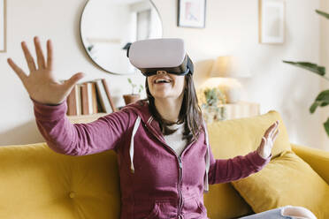 Happy young woman wearing virtual reality headset gesturing at home - XLGF02616