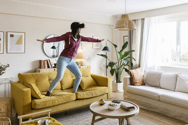 Young woman wearing virtual reality headset standing with arms outstretched on sofa in living room - XLGF02618