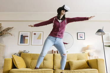 Happy woman wearing virtual reality headset standing with arms outstretched on sofa - XLGF02619