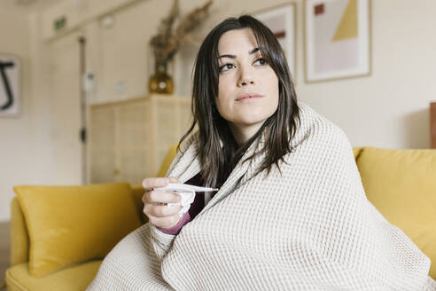 Beautiful woman wrapped in blanket holding thermometer in living room - XLGF02624