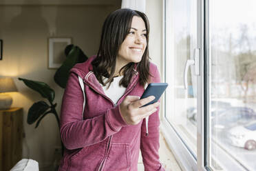 Glückliche schöne Frau mit Smartphone schaut durch das Fenster - XLGF02636