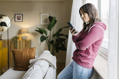 Frau schickt Textnachrichten über ihr Smartphone am Fenster zu Hause - XLGF02637