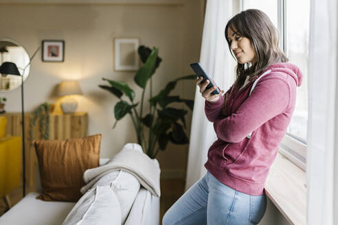 Woman text messaging through smart phone by window at home - XLGF02637