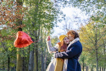 Young man kissing girlfriend holding red heart shape balloon at park - EIF03266