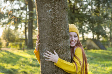 Smiling redhead woman hugging tree at park - EIF03292