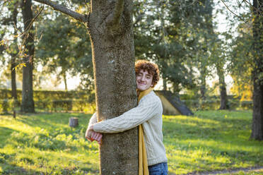 Lächelnder junger Mann, der einen Baum im Park umarmt - EIF03293