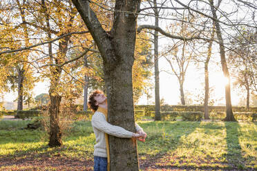 Junger Mann umarmt einen Baum und blickt auf einen herbstlichen Park - EIF03294