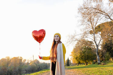 Smiling redhead woman holding red heart shape balloon in autumn park - EIF03298