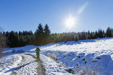 Die Sonne scheint über einem älteren Wanderer, der auf einer verschneiten Straße im Naturschutzgebiet Perlenbach-Fuhrtsbachtal wandert - GWF07301