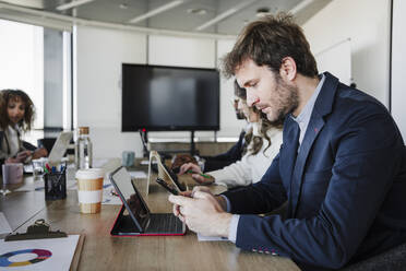 Geschäftsmann, der ein Smartphone benutzt, sitzt neben Kollegen am Schreibtisch im Büro - EBBF05273