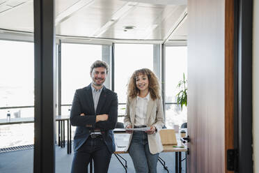 Smiling businessman with arms crossed by colleague holding document in office - EBBF05330