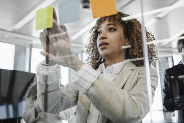 Young businesswoman with brown curly hair sticking adhesive notes on glass wall in office - EBBF05339