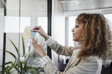 Businesswoman with curly hair sticking adhesive notes on glass wall in office - EBBF05340