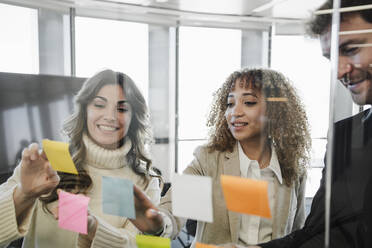 Businessman and businesswomen sticking adhesive notes seen through glass wall - EBBF05346