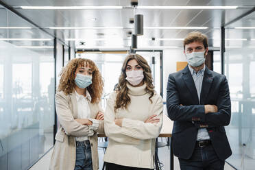 Geschäftsmann und Geschäftsfrau mit Gesichtsschutzmaske stehen mit verschränkten Armen in einem Büro - EBBF05349