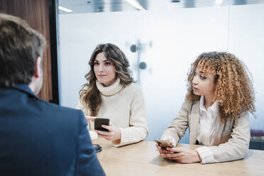 Geschäftsfrauen mit Smartphones im Gespräch mit einem Geschäftsmann, der am Schreibtisch sitzt - EBBF05350