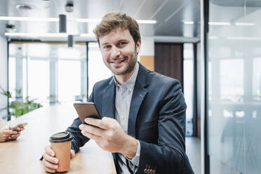 Smiling businessman with disposable coffee cup and smart phone sitting at desk - EBBF05355