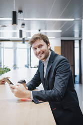 Happy businessman with mobile phone and disposable coffee cup sitting at desk in office - EBBF05356