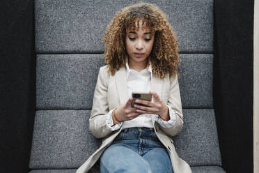 Junge Geschäftsfrau mit braunen lockigen Haaren, die mit ihrem Smartphone im Büro Textnachrichten verschickt - EBBF05362