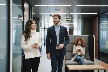 Businesswoman using smart phone by colleagues walking in corridor - EBBF05364