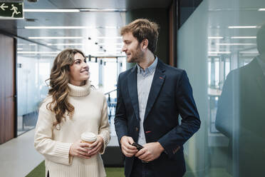 Smiling businessman and businesswoman looking at each other by glass wall in office - EBBF05367