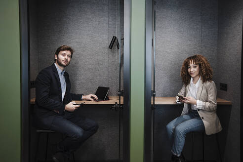 Geschäftsfrau und Geschäftsmann mit Mobiltelefonen sitzen am Schreibtisch in einer Kabine - EBBF05372