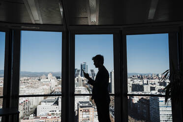 Silhouette eines Geschäftsmannes, der mit seinem Smartphone an einer Glaswand im Büro Textnachrichten verschickt - EBBF05382