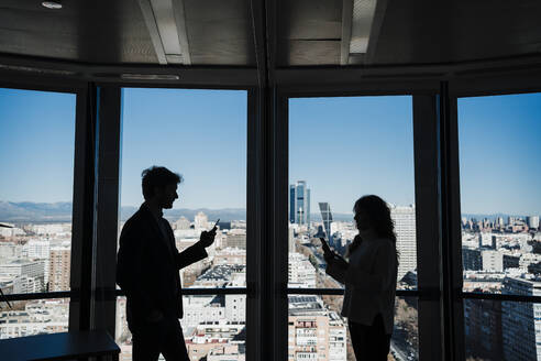 Silhouette eines Geschäftsmannes und einer Geschäftsfrau, die Smartphones an einer Glaswand im Büro benutzen - EBBF05384