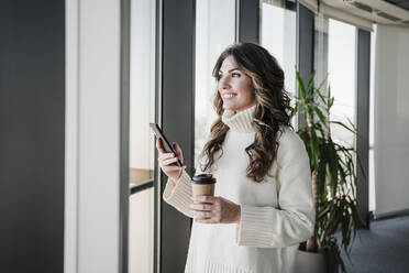 Lächelnde Geschäftsfrau mit Smartphone hält Einwegbecher in der Nähe des Fensters im Büro - EBBF05390